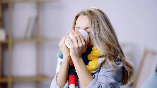 Femme blonde malade en écharpe colorée tenant tasse tout en buvant du thé à la maison — Photo de stock