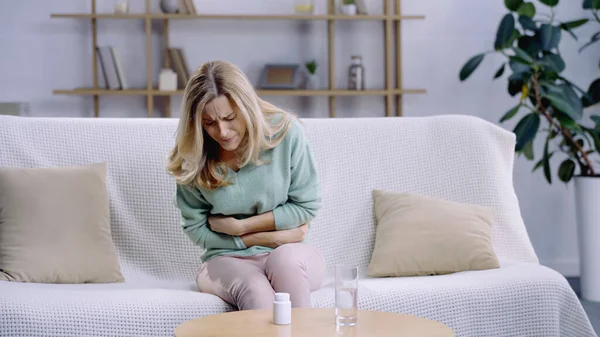 Blondine mit Bauchschmerzen, während sie neben Glas Wasser und Tabletten auf dem Couchtisch sitzt — Stockfoto