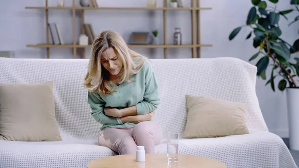 Mujer rubia con dolor abdominal mientras está sentada cerca de un vaso de agua y pastillas en la mesa de café — Stock Photo