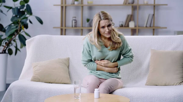 Frau hat Bauchschmerzen, während sie neben Glas Wasser und Tabletten auf dem Couchtisch sitzt — Stockfoto