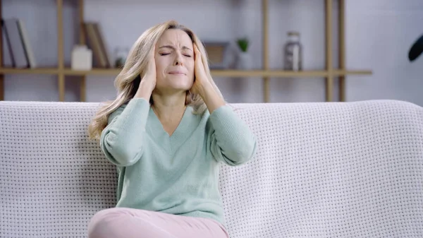 Blondine mit geschlossenen Augen hat Migräne, während sie im Wohnzimmer den Kopf berührt — Stockfoto