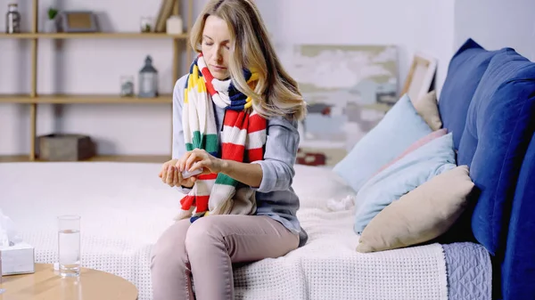 Sick woman in colorful scarf sitting on bed and taking pills — Stock Photo