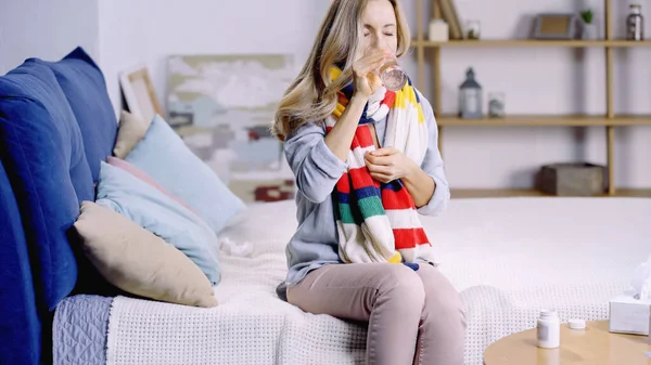 Больная женщина в красочном шарфе сидит на кровати и пьет воду, принимая таблетки — стоковое фото