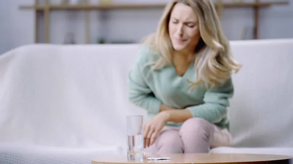 Mujer rubia que sufre de dolor de período cerca de vaso de agua y botella con pastillas en la mesa de café — Stock Photo