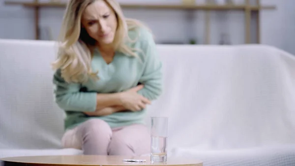 Mulher loira sofrendo de dor de estômago perto de vidro de água e garrafa com pílulas na mesa de café — Fotografia de Stock