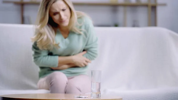 Mujer rubia que sufre de dolor de estómago cerca de vaso de agua y botella con pastillas en la mesa de café — Stock Photo