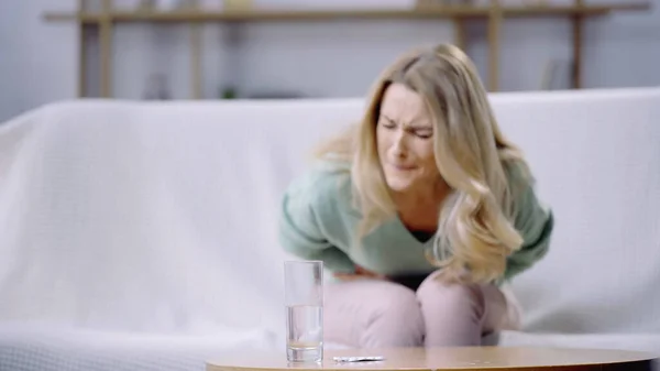 Mujer que sufre de dolor de período cerca de vaso de agua y botella con pastillas en la mesa de café — Stock Photo