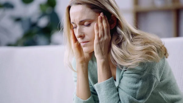 Traurige Frau mit geschlossenen Augen mit Migräne und Berührung des Kopfes im Wohnzimmer — Stockfoto