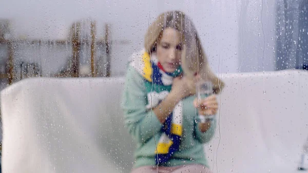 Sick woman in colorful scarf holding glass of water behind wet window with rain drops — Stock Photo