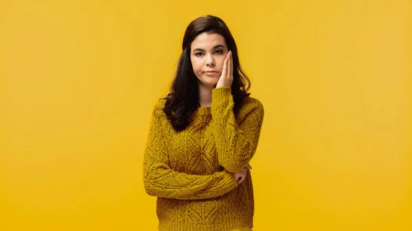 Displeased brunette woman in sweater touching cheek isolated on yellow — Stock Photo