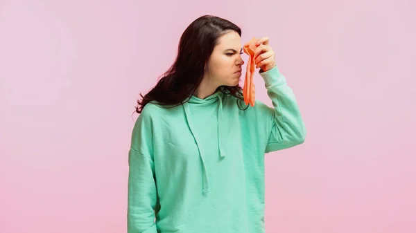 Young and brunette woman in turquoise hoodie smelling stinky socks isolated on pink — Stock Photo