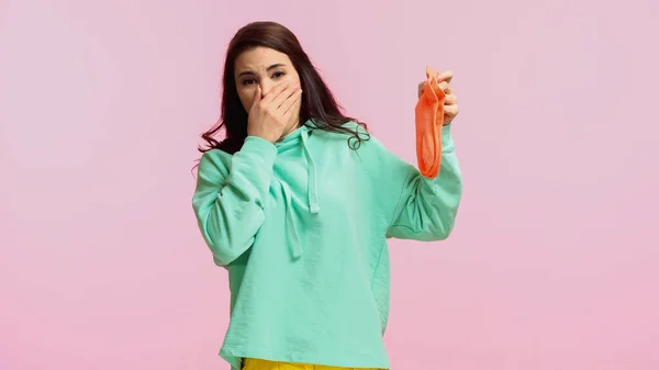 Young and brunette woman in turquoise hoodie plugging nose while holding stinky socks isolated on pink — Stock Photo