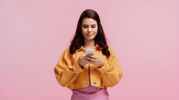 Sonriente mujer joven en chaqueta naranja mensajes de texto en el teléfono inteligente aislado en rosa - foto de stock