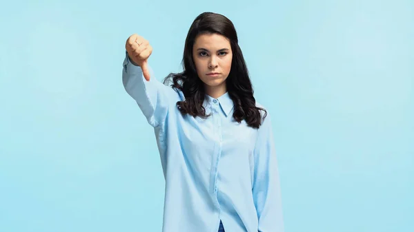 Unpleased young woman in shirt standing and showing thumb down isolated on blue — стоковое фото