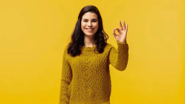 Mujer feliz en suéter mirando a la cámara mientras muestra signo ok aislado en amarillo - foto de stock