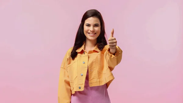 Счастливая молодая женщина в оранжевой джинсовой куртке показывает большой палец вверх изолирован на розовый — стоковое фото