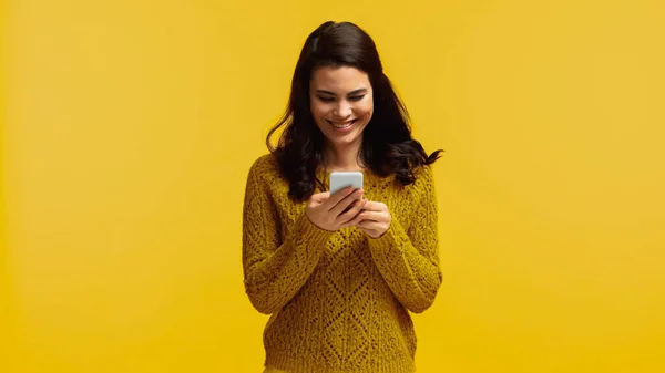 Cheerful and brunette woman in sweater using smartphone isolated on yellow — Stock Photo