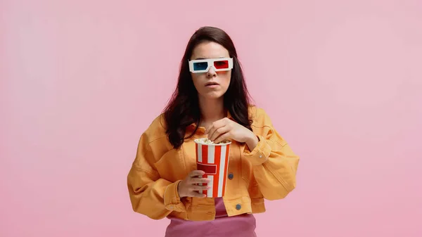 Morena joven en chaqueta de mezclilla naranja y gafas 3d sosteniendo palomitas de maíz cubo aislado en rosa - foto de stock