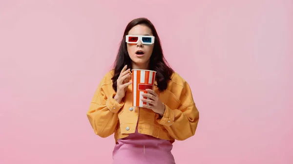 Mujer joven asustada en chaqueta de mezclilla naranja y gafas 3d sosteniendo palomitas de maíz cubo aislado en rosa - foto de stock