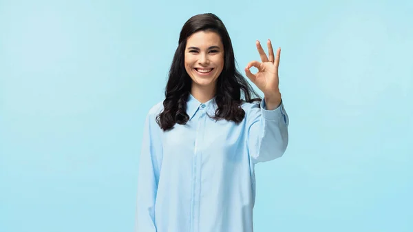 Freudige junge Frau im Hemd blickt in die Kamera, während sie ok isoliert auf blau zeigt — Stockfoto