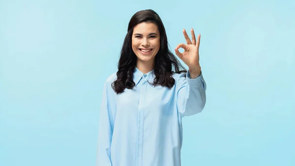 Alegre joven mujer en camisa mirando a la cámara mientras que muestra ok aislado en azul - foto de stock