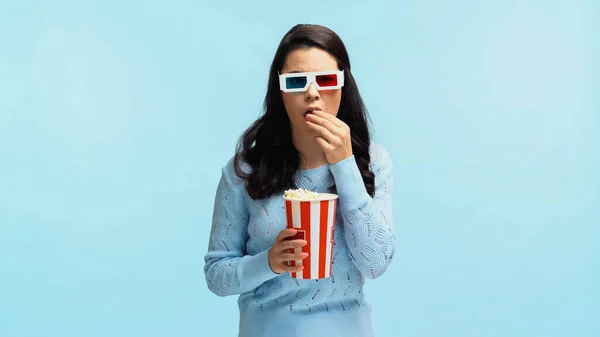 Morena mujer joven en gafas 3d comer palomitas de maíz y ver la película aislado en azul - foto de stock