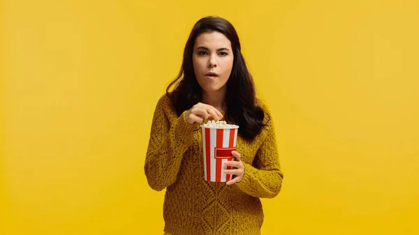 Сфокусированная брюнетка в свитере ест попкорн и смотрит фильм, изолированный на желтом — стоковое фото