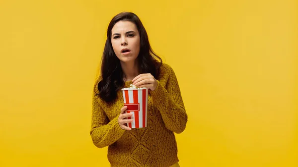 Сфокусированная молодая женщина в свитере ест попкорн и смотрит фильм изолирован на желтый — стоковое фото