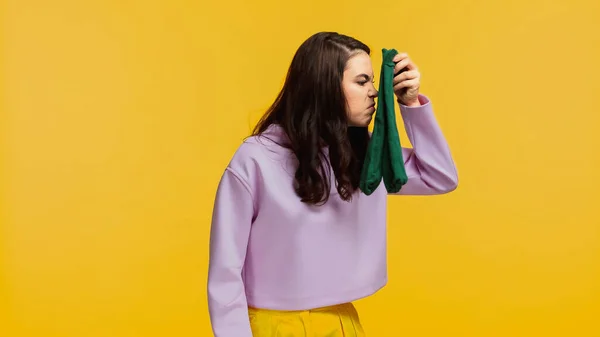 Brunette woman in purple sweatshirt smelling stinky socks isolated on yellow — Stock Photo