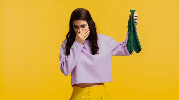 Brunette woman in purple sweatshirt plugging nose and holding stinky socks isolated on yellow — Stock Photo