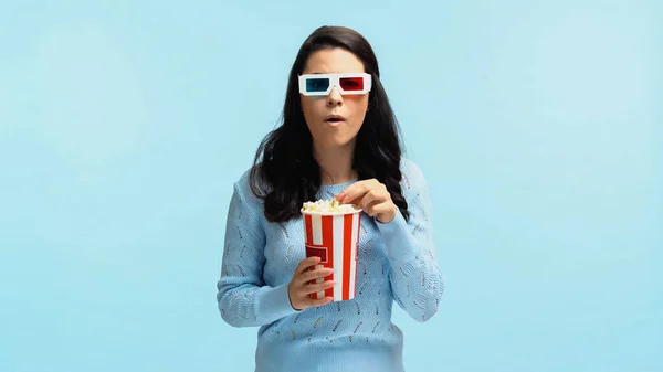 Шокована молода жінка в 3d окулярах їсть попкорн під час перегляду фільму ізольовано на синьому — стокове фото