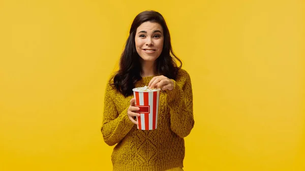 Happy young woman in sweater holding popcorn and watching comedy movie isolated on yellow — Stock Photo