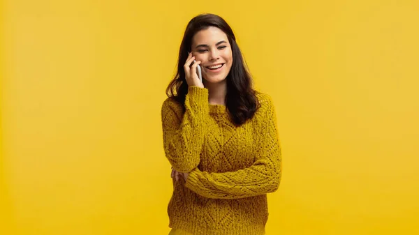 Mujer alegre y morena en suéter hablando en smartphone aislado en amarillo - foto de stock