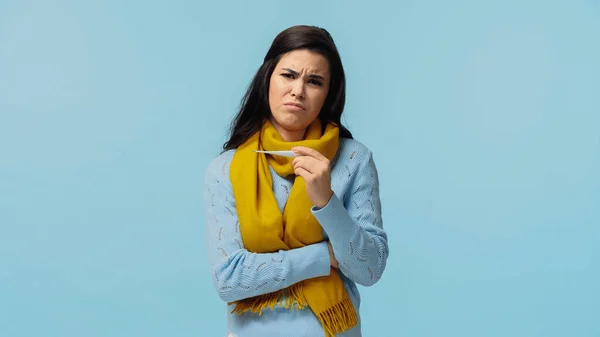 Засмучена і хвора жінка в шарфі тримає електронний термометр ізольовано на синьому — стокове фото