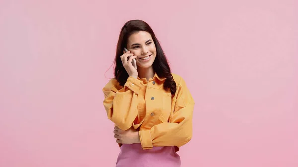 Mujer joven feliz en chaqueta de mezclilla naranja hablando en el teléfono inteligente aislado en rosa - foto de stock