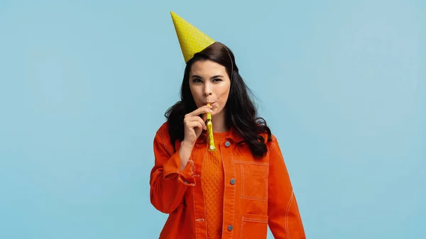 Щаслива молода жінка в помаранчевій куртці і кепці, що дме партійний ріг ізольовано на синьому — стокове фото