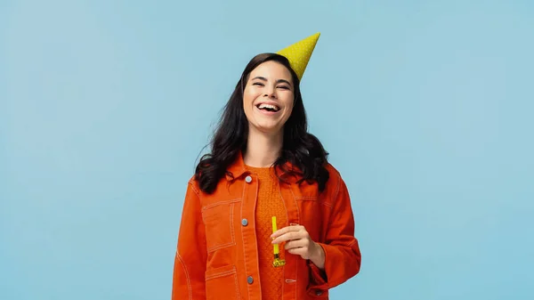 Excited young woman in orange jacket and festive cap holding party horn while laughing isolated on blue — Stock Photo