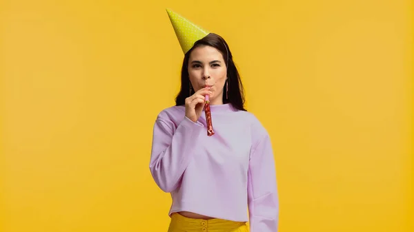 Alegre joven mujer en púrpura sudadera y partido gorra soplado cuerno aislado en amarillo - foto de stock