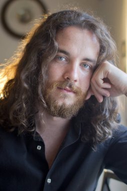 Man with gray eyes, beard and long brown hair. Smiling model with head resting on hand.