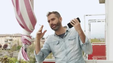 İtalyan adam akıllı telefonuyla bir sesli mesaj dinler. Çekici adam bulutlu bir günde balkonda sigara içiyor. Covid karantinası sırasında iletişim ve sosyal izolasyon.