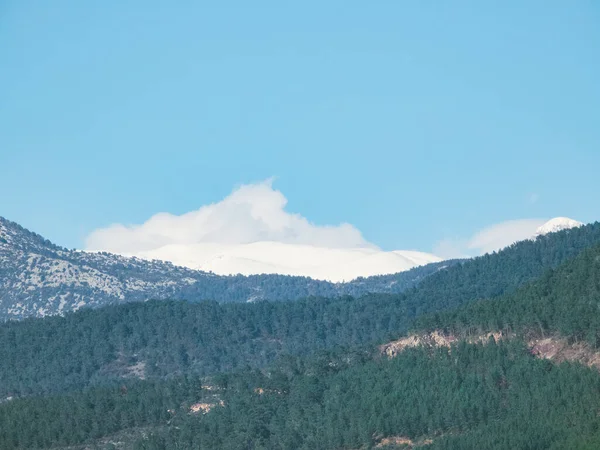 Snow-capped mountain peak with a cap of clouds, surrounded by green mountain trees, on a sunny day