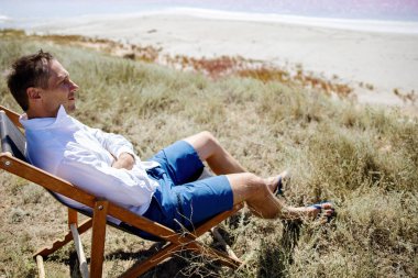 Güneşli bir günde, pembe denizin kumsalında güverte koltuğunda oturan bir iş adamı..