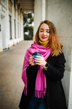 Şık, mutlu genç bir bayan pembe bir eşarp takıyor, elinde bir kahve tutuyor. Sonbahar şehrinde gülümseyen bir kızın portresi.