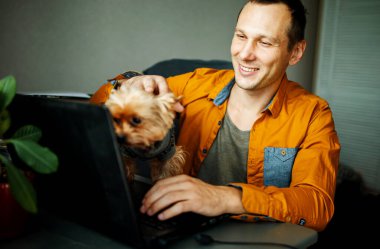 Evde köpeğiyle dizüstü bilgisayar kullanan yakışıklı gülen adam..