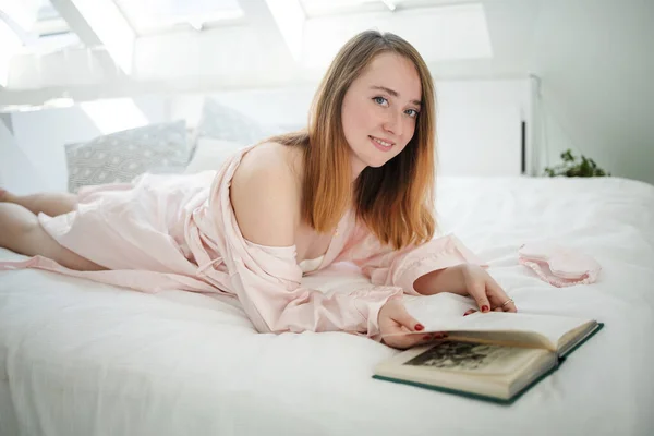 光の中で窓の近くのベッドの上に寝そべっているパジャマ姿のリラックスした白人の少女 彼女は本で晴れた春の朝を楽しんでいる — ストック写真