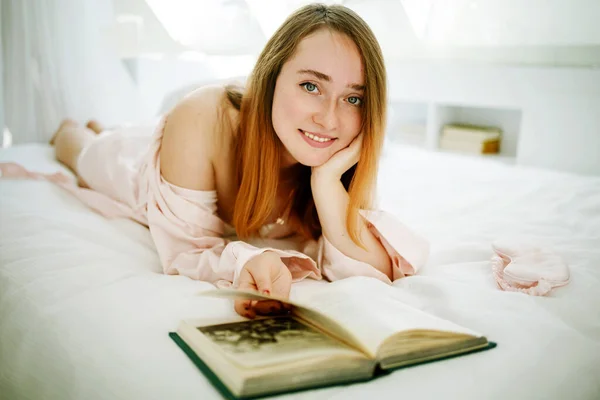 光の中で窓の近くのベッドの上に寝そべっているパジャマ姿のリラックスした白人の少女 彼女は本で晴れた春の朝を楽しんでいる — ストック写真
