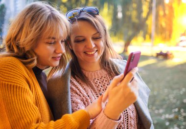Anne ve kızı, sonbahar parkında sarı giyinmiş bir şekilde telefon kullanıyorlar.