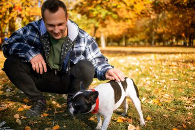 Sonbahar parkında çimlerin üzerinde köpeğini kucaklayan bir adam.