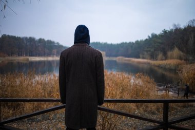 Genç adam tek başına göl köprüsünde duruyor ve gri bulutlu gökyüzünde güneşin doğuşuna bakıyor. Suyun üzerinde sis. Sisli hava. Sabahın erken saatlerinde. Karanlık, korkunç bir an ve kasvetli bir atmosfer. Arka plan.
