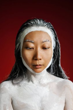 Yüzünde kil maske var, yumuşaklığı seviyor, evde güzellik tedavileri uyguluyor, yarı çıplak poz veriyor, kırmızı arka planda izole edilmiş. Cilt bakımı kavramı.